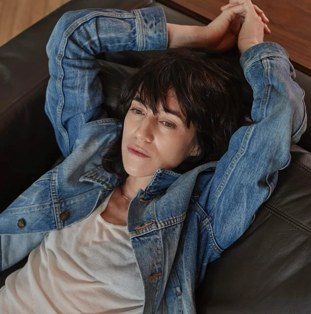 Adult woman in a blue denim jacket poses while lying down on a dark brown leather couch in an indoor setting. Casual and relaxed.