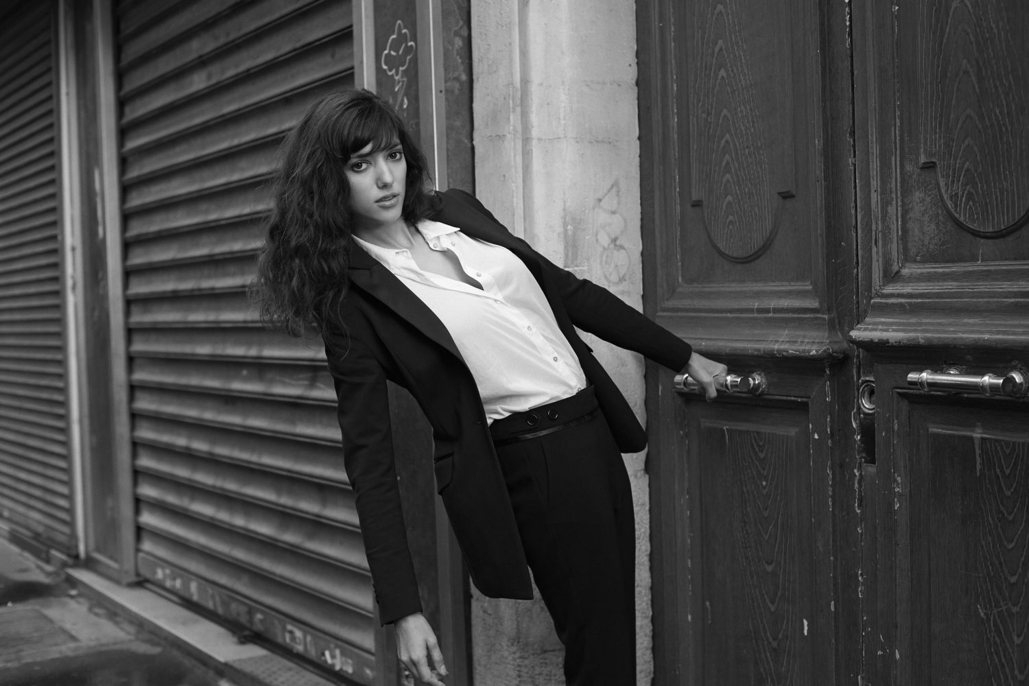 Black and white portrait of a young woman in a suit standing in front of an old door and corrugated metal wall on a city street.
