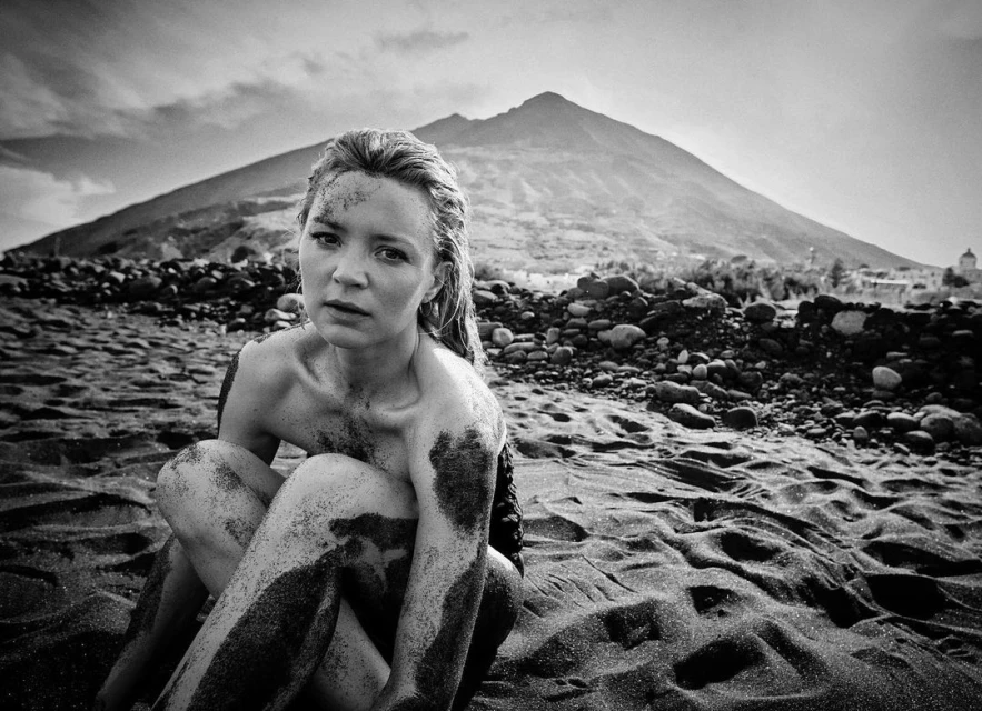 Black and white shot of a woman sitting on a black sand beach with a volcanic mountain in the background, monochrome scenic view.
