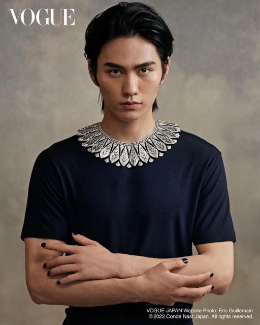 Asian man with dark hair wearing black T-shirt and silver neck piece, posing in a Vogue Japan portrait against neutral background.