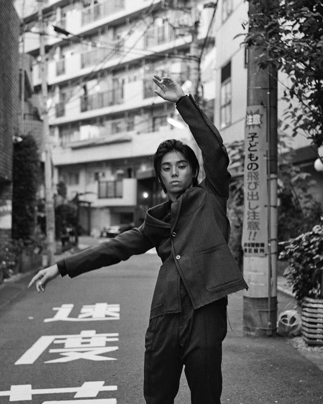 Monochrome portrait of a person in a jacket and trousers posing in a street framed by buildings and a utility pole, with graffiti on the ground.