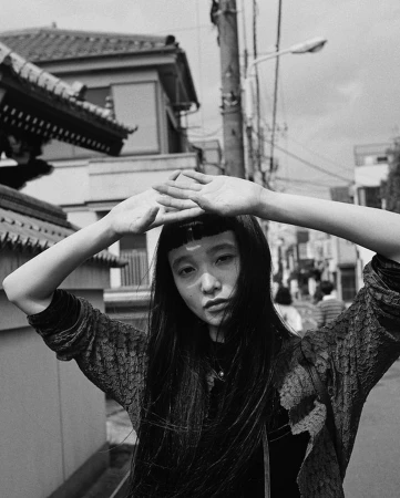 Black and white portrait of a woman with long dark hair and bangs holding her hands above her head on a street.