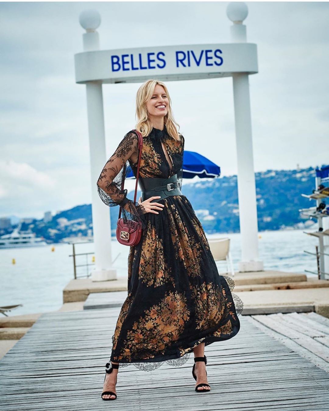 A blonde woman in a long, floral dress stands on a wooden dock with a small purse and high-heeled sandals with the ocean behind her.
