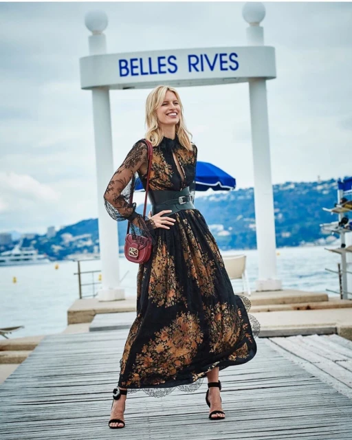 A blonde woman in a long, floral dress stands on a wooden dock with a small purse and high-heeled sandals with the ocean behind her.