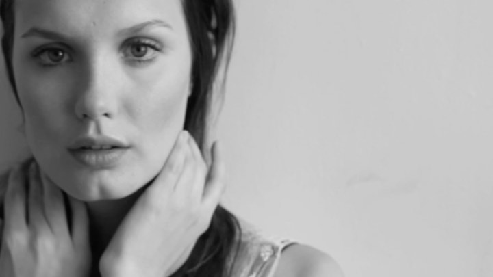 Monochrome portrait of a fair-skinned woman with hands near her neck against a plain background, focusing on her face and expression.