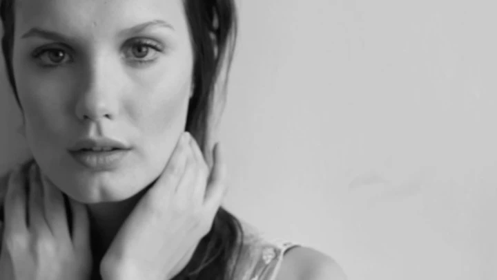 Monochrome portrait of a fair-skinned woman with hands near her neck against a plain background, focusing on her face and expression.