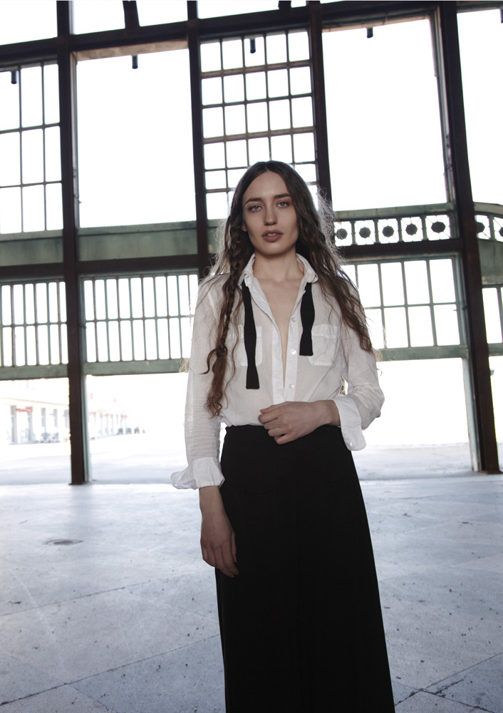 Portrait of a woman with long hair wearing a white shirt and black skirt in a spacious indoor location with large windows.