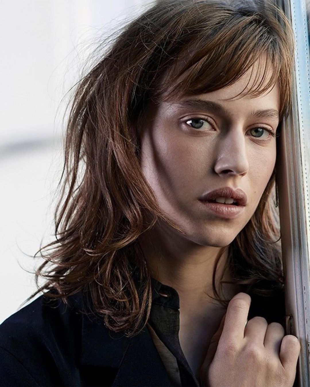 Close-up portrait of a young woman with brown hair and bangs leaning against a window frame wearing a black jacket