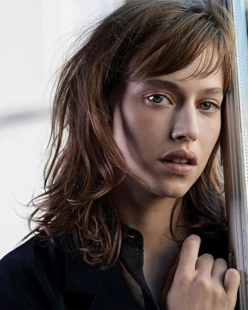 Close-up portrait of a young woman with brown hair and bangs leaning against a window frame wearing a black jacket
