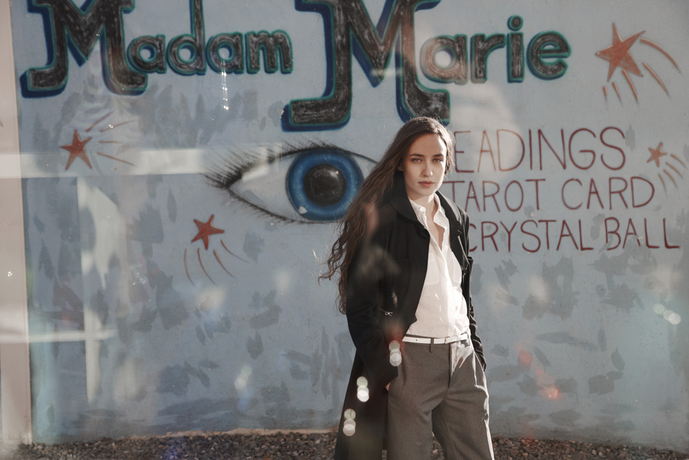 Adult female in trench coat stands in front of a store window with tarot card and crystal ball signage, gazing directly at the camera.