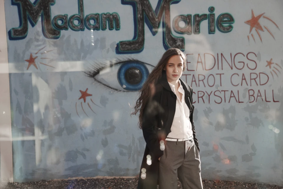 Adult female in trench coat stands in front of a store window with tarot card and crystal ball signage, gazing directly at the camera.