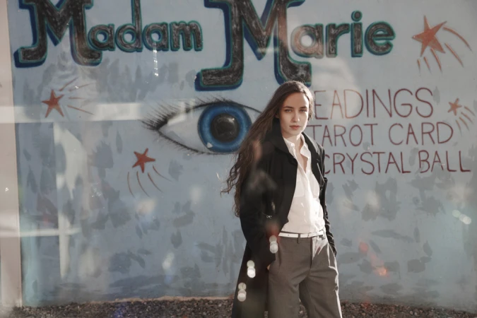 Adult female in trench coat stands in front of a store window with tarot card and crystal ball signage, gazing directly at the camera.