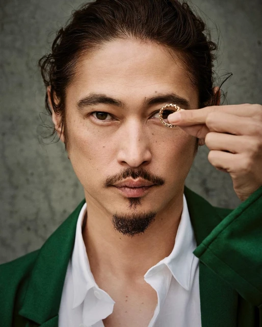 Portrait of an adult man with dark hair and a goatee, wearing a green jacket and holding a gold ring up to his eye.