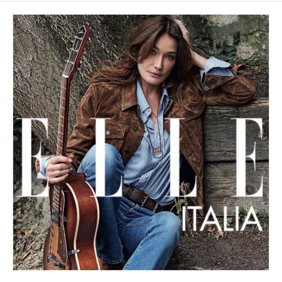 Carla Bruni poses with a guitar for Elle Italia. She wears a brown jacket, blue jeans, and a light blue shirt with a necklace. The background is a forest.