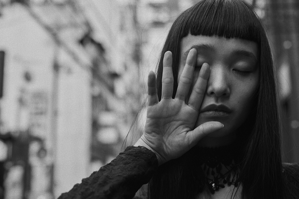 Black and white portrait of a young Asian woman with bangs shielding her eye with her hand against an out-of-focus urban backdrop.