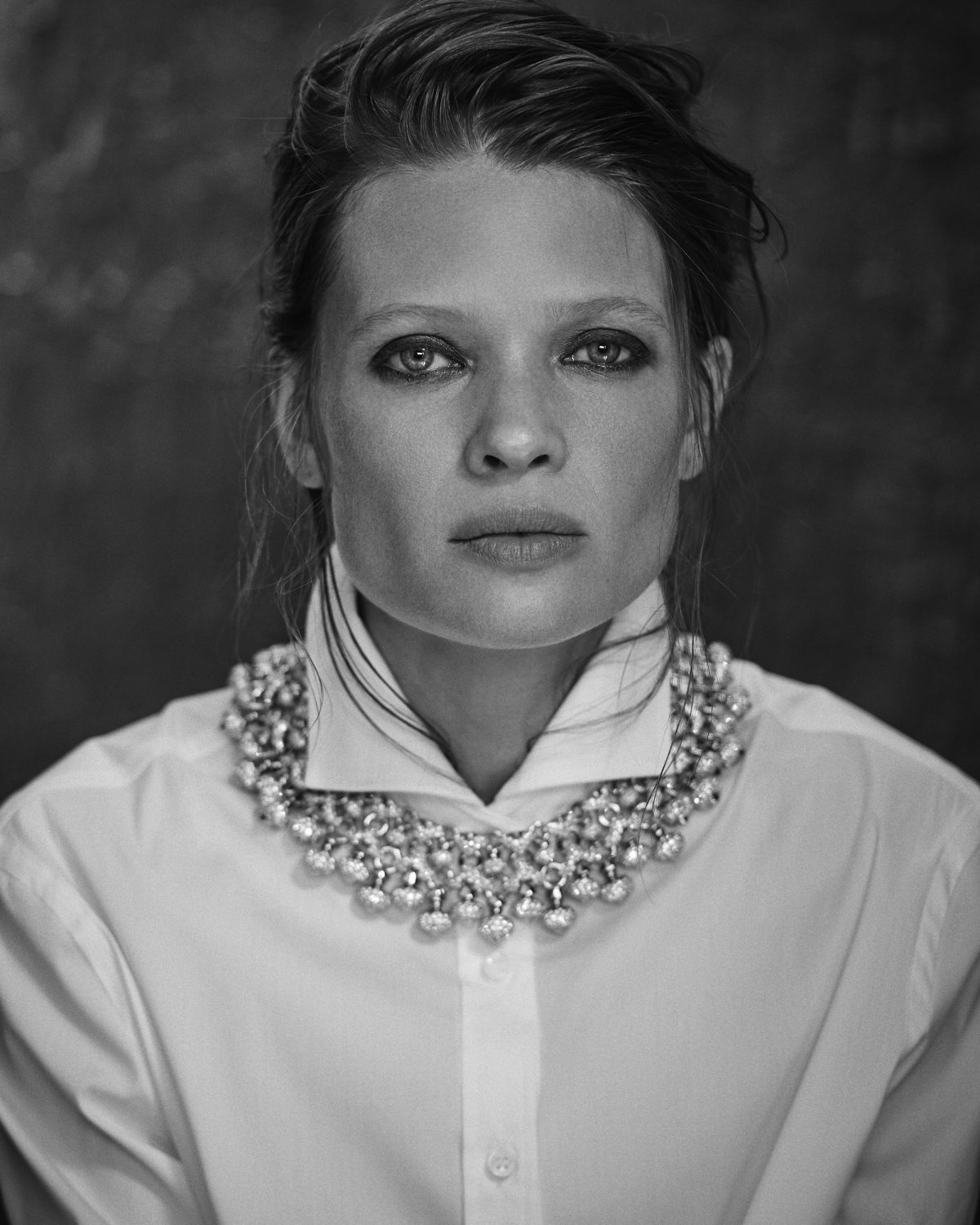 Monochrome portrait of a fair-skinned adult female with striking eye makeup wearing a white collared shirt and a statement necklace.