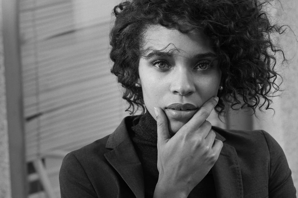 Black and white portrait of a woman with curly hair wearing a blazer, looking thoughtfully at the camera with hand on chin.