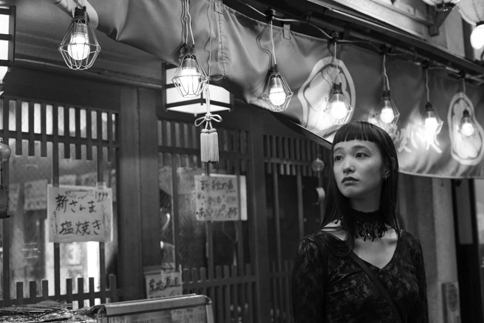 Monochrome portrait of a young Asian woman in black lace top and choker, gazing left, with vintage storefront and hanging lights.