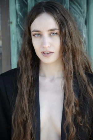 Portrait of a woman with long wavy brown hair wearing a black jacket against a green wooden door.