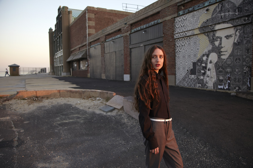 Young woman with long brown hair poses outdoors near a brick building with mural art, wearing a dark shirt and grey pants with a white belt.