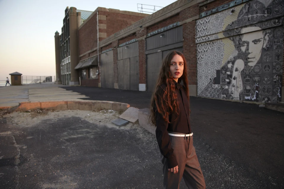 Young woman with long brown hair poses outdoors near a brick building with mural art, wearing a dark shirt and grey pants with a white belt.