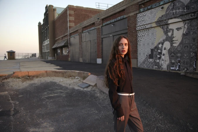 Young woman with long brown hair poses outdoors near a brick building with mural art, wearing a dark shirt and grey pants with a white belt.