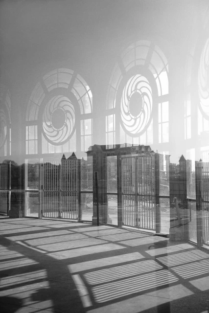 Black and white photograph of sunlight streaming across a floor with shadows, framed by large arched windows and a distant city skyline.