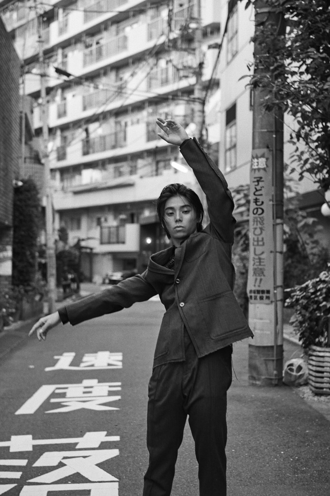 Monochrome portrait of a fashion model in an alleyway in a suit, posing with arms outstretched against urban street with Japanese text.