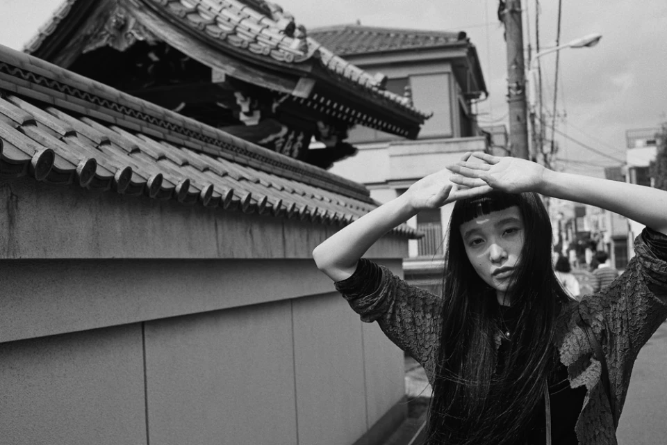 Monochrome portrait of a young Asian adult woman posing outdoors with a temple roof and residential buildings in the background.