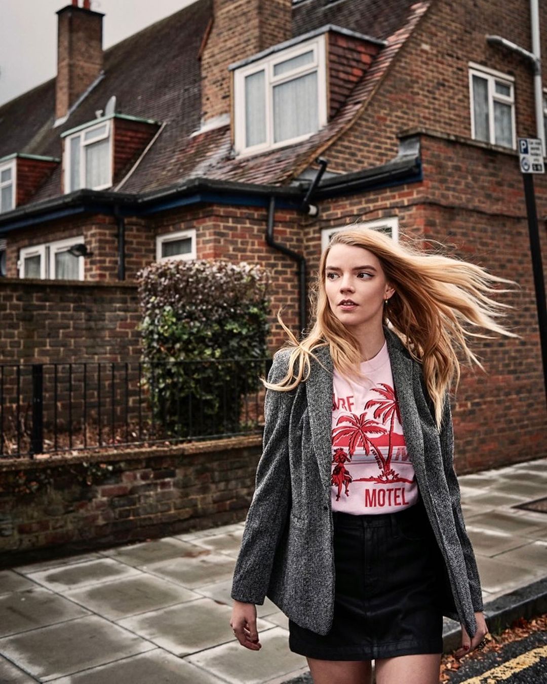 Anya Taylor-Joy on a city street wearing a blazer, T-shirt with palm trees and a mini skirt, posing against brick buildings.