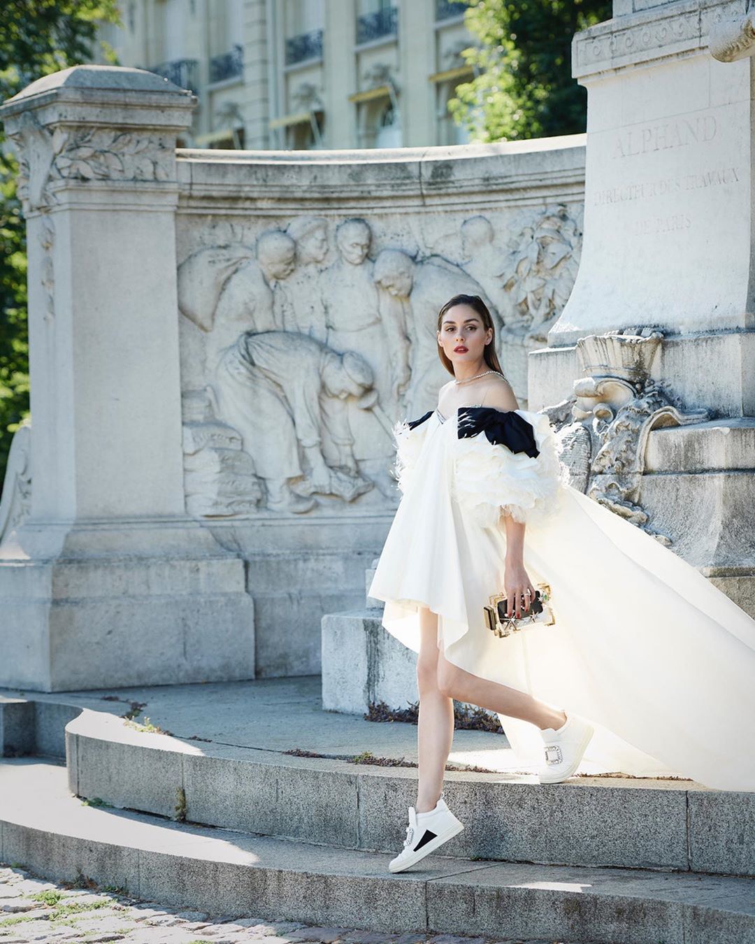 Elegant woman in a white dress and black accents poses on stone steps in front of a monument with bas-relief sculptures outdoors.