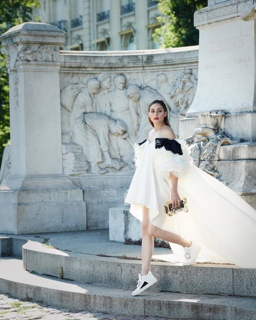 Elegant woman in a white dress and black accents poses on stone steps in front of a monument with bas-relief sculptures outdoors.