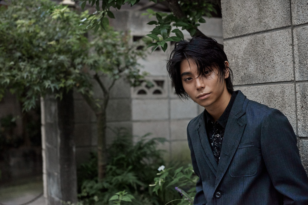 Portrait of an Asian adult male in a dark blue suit, standing outdoors against a textured concrete wall with green plants and foliage.
