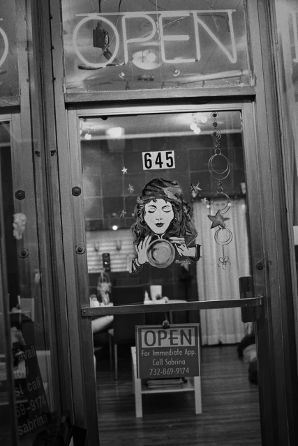 Black and white photo of a storefront window with an 'Open' sign, a graphic of a woman, and a glimpse into the shop's interior.