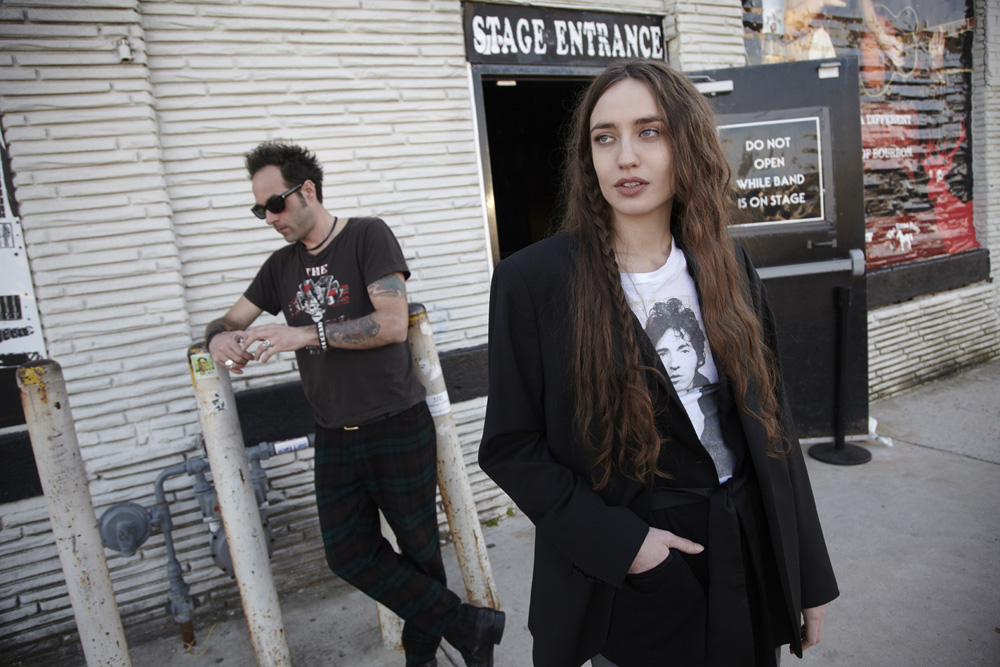 Two people stand outside a brick building next to a black door labeled 'Stage Entrance'; the woman wears a black blazer and the man leans on a metal barrier.