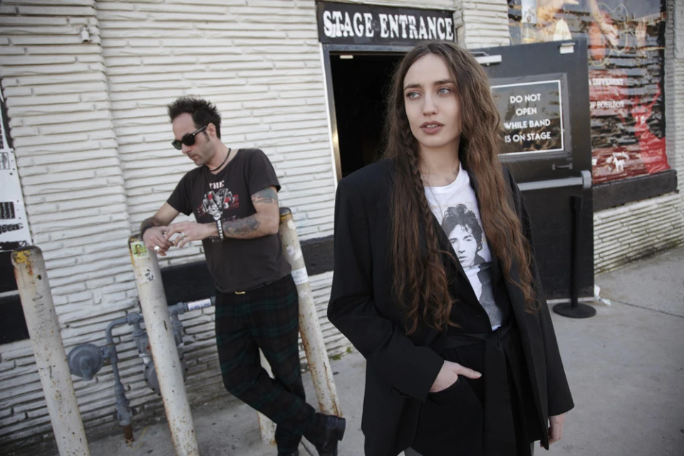 Two people stand outside a brick building next to a black door labeled 'Stage Entrance'; the woman wears a black blazer and the man leans on a metal barrier.