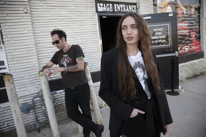 Two people stand outside a brick building next to a black door labeled 'Stage Entrance'; the woman wears a black blazer and the man leans on a metal barrier.