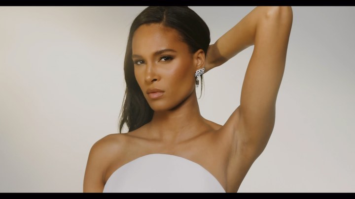 Close-up shot of a beautiful dark-skinned woman with long dark hair, posing in a strapless top with a hand behind her head, indoors.