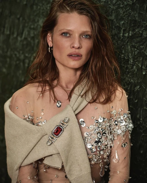 Close-up shot of a woman with red hair wearing a sheer embellished top and draped knitwear, standing against a mottled green background.