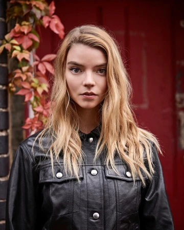 Portrait of Anya Taylor-Joy wearing a black leather jacket against a dark red wooden door with autumn foliage.