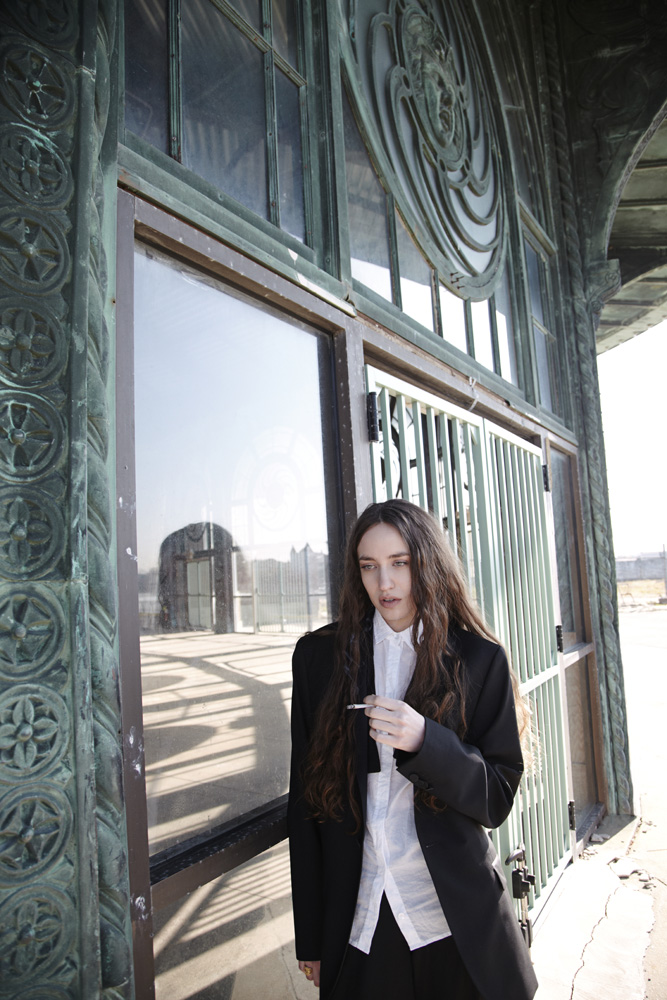 Woman in black blazer and white shirt standing against weathered metallic architecture with ornate patterns.