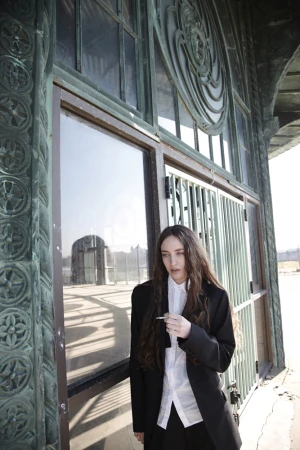 Woman in black blazer and white shirt standing against weathered metallic architecture with ornate patterns.