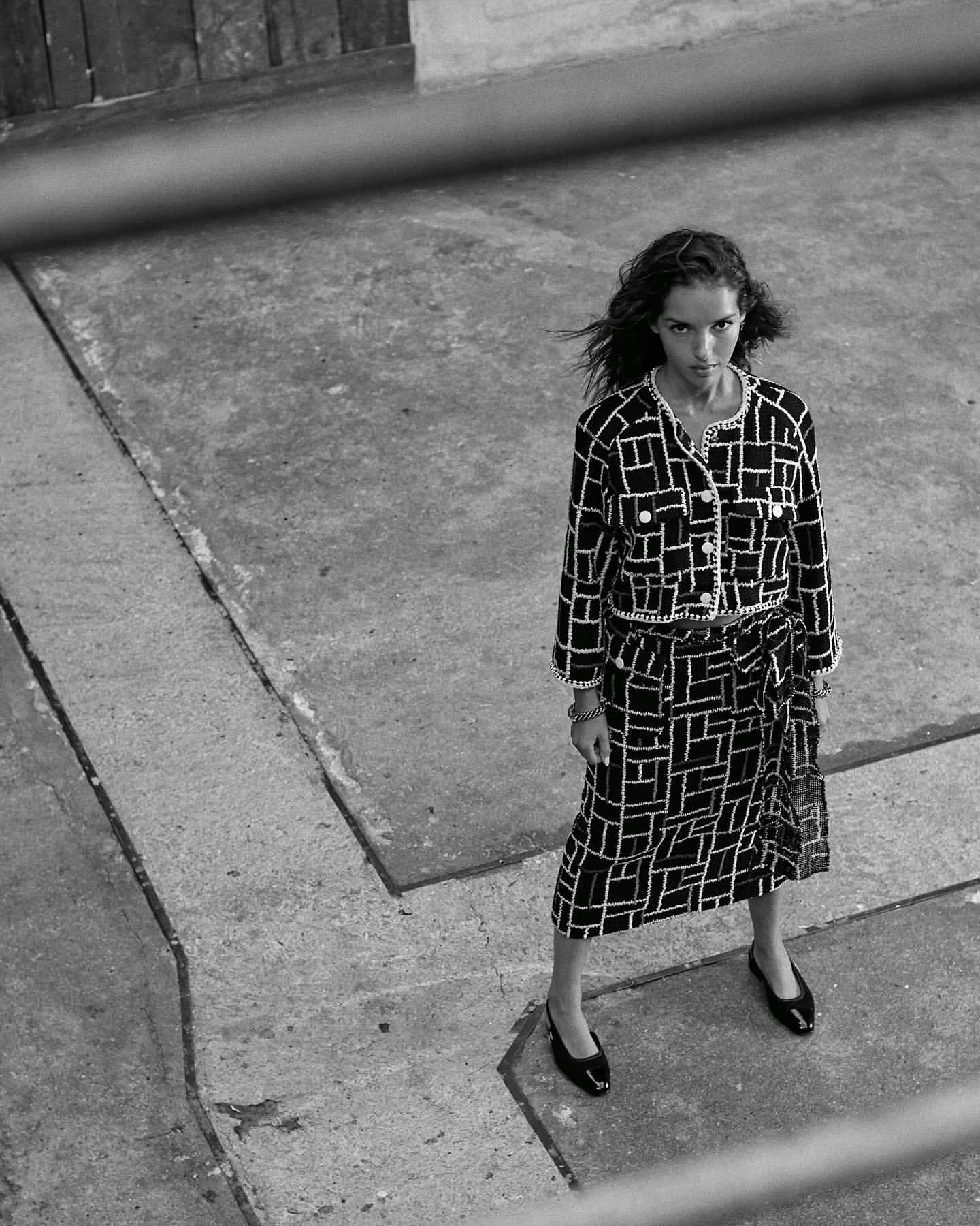 Woman in a black and white patterned two-piece outfit stands on a concrete surface in a fashion editorial style photograph.