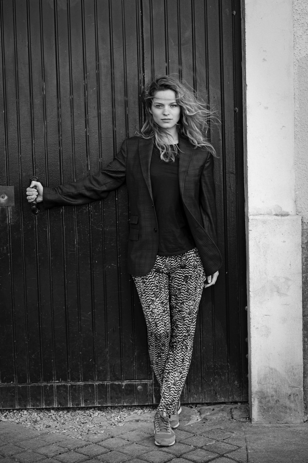Black and white portrait of a woman leaning against a dark wooden door wearing a blazer and patterned trousers by Eric Guillemain.