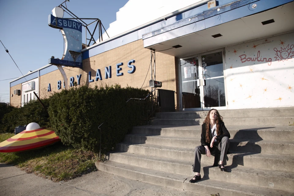 A young woman poses on concrete steps in front of a vintage building with faded signage and retro architectural details on a sunny day.