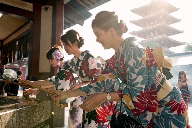 Three women in traditional floral kimonos holding wooden ladles at a water purification fountain in Japan.