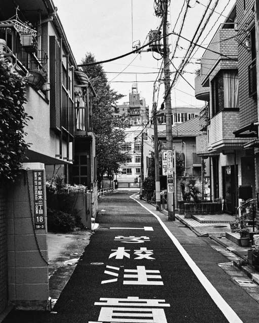 Monochrome view of a narrow city street in Japan with power lines above and Japanese text painted on the asphalt street surface.