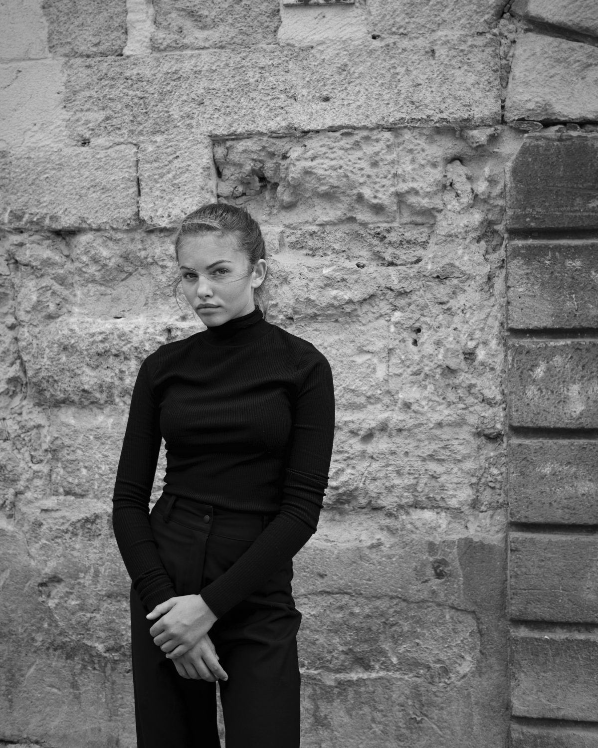 Monochrome portrait of a young woman in a black turtleneck standing against an ancient stone wall