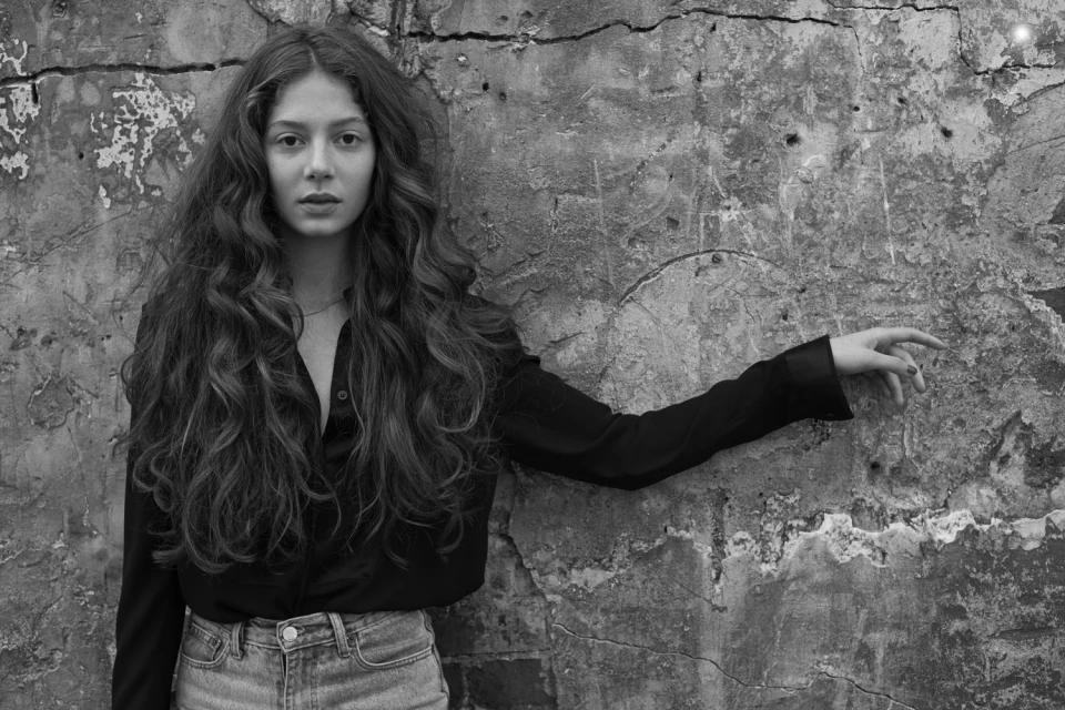 Portrait of an adult female with long wavy hair posing in front of a cracked concrete wall wearing a dark-colored button-down shirt and denim jeans.