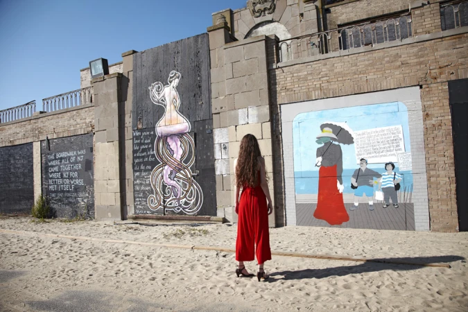 Woman in red jumpsuit stands on beach facing brick wall with mural art featuring figures and text, under a clear blue sky.
